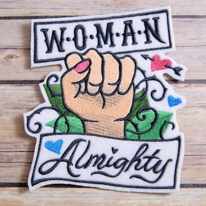 May include: Embroidered patch featuring a raised fist, green vines, and a banner that reads "WOMAN Almighty". The patch has a white background with black stitching and includes a pink heart with an arrow and a blue heart.