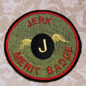 May include: A green and red circular patch with the text "JERK MERIT BADGE" in red embroidery. A black circle with a gold "J" inside is centered on the patch with gold wings on either side.