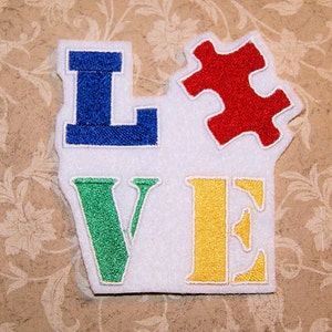 May include: A white felt patch with the word "LOVE" embroidered in different colors. The "L" is blue, the "V" is green, the "E" is yellow, and a red puzzle piece is above the word.