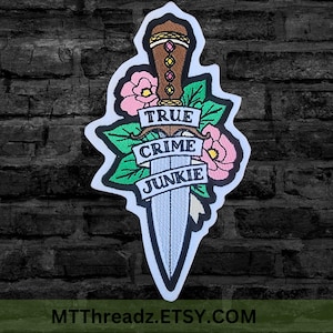 May include: An embroidered patch featuring a brown dagger with a jeweled hilt, surrounded by pink flowers and green leaves. A white banner draped across the blade displays the black text TRUE CRIME JUNKIE in a bold, capitalized font style.