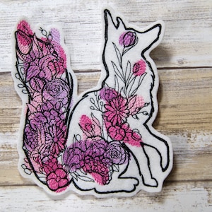 May include: A white felt applique of a fox with pink and purple floral embroidery. The fox is sitting with its tail curled around its body.