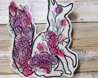 May include: A white felt applique of a fox with pink and purple floral embroidery. The fox is sitting with its tail curled around its body.