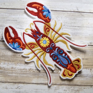 May include: A white felt embroidered patch of a red lobster with yellow, blue, and orange floral details.