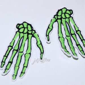 May include: Two green skeleton hand iron-on patches. The patches are detailed with white stitching and are perfect for adding a spooky touch to clothing or accessories.