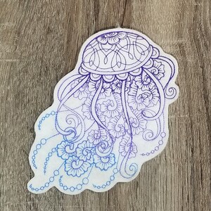 Peut inclure: Un patch en tissu blanc avec un motif brodé d'une méduse violette et bleue aux détails floraux complexes.