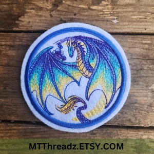 May include: A circular embroidered patch featuring a dragon with blue, teal, and gold thread. The dragon is depicted in a coiled position with wings spread against a white background, enclosed by a blue border. The text MTTTHREADZ.ETSY.COM is visible.