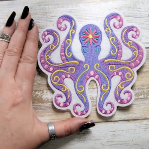 May include: Purple and yellow embroidered octopus with pink accents on a white felt background. The octopus has a flower on its head.