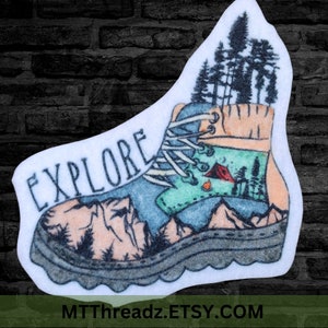 May include: A white sticker with a blue and green hiking boot with a mountain and forest scene inside. The boot is surrounded by mountains and trees. The word "EXPLORE" is written in black on the sticker.