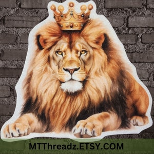 May include: A golden crown sits atop the head of a majestic lion with a thick mane. The lion has a serious expression and is looking directly at the viewer. The background is a brick wall.