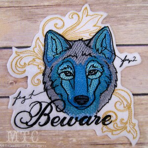 May include: Embroidered patch featuring a blue and gray wolf's head with the word "Beware" in a decorative script below. The wolf's head is surrounded by gold floral designs.