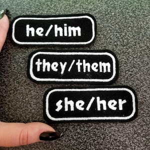 May include: Three black fabric patches with white embroidered text. The patches read "he/him", "they/them", and "she/her".