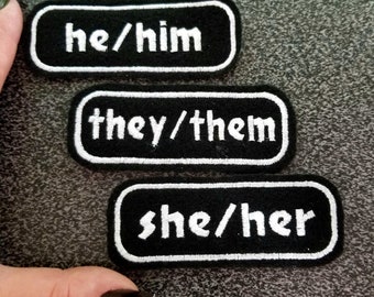 May include: Three black fabric patches with white embroidered text. The patches read "he/him", "they/them", and "she/her".