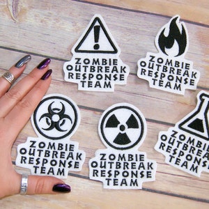 May include: Six white embroidered patches with black text and symbols. The patches read "Zombie Outbreak Response Team" and feature symbols like a biohazard symbol, a radiation symbol, a fire symbol, a warning symbol, and a beaker.