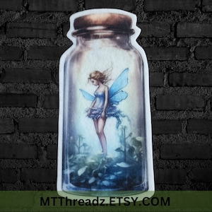 May include: A glass jar with a cork lid contains a blue fairy with wings. The fairy is standing on a bed of green leaves and flowers. The jar is clear and has a textured surface.