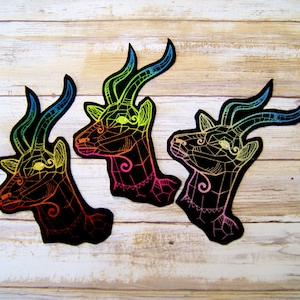 May include: Three black iron-on patches with a rainbow outline of an antelope head. The patches are on a light wood background.