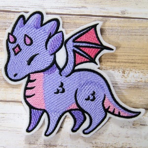 May include: Purple and pink embroidered dragon patch with a red wing. The dragon has a cute cartoon style and is standing on all fours.