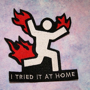 May include: A black and white embroidered patch with a stick figure on fire. The text "I TRIED IT AT HOME" is below the figure.