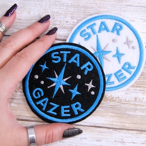 May include: Two embroidered iron-on patches with blue trim. One patch is white with the text "STAR GAZER" in blue and silver stars. The other patch is black with the text "STAR GAZER" in blue and silver stars.