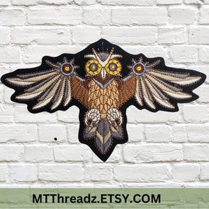 May include: A brown and gray embroidered owl patch with yellow eyes. The owl is spread out with its wings open. The patch is on a white brick background.