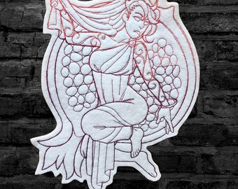 May include: Embroidered patch featuring a woman with horns and flowing hair, sitting inside a pomegranate. The design is outlined in white and filled with a pink-red color.