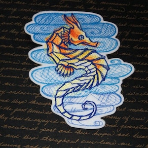 Peut inclure: Un patch brodé blanc représentant un hippocampe jaune et orange avec des nuages bleus et blancs. L'hippocampe est détaillé avec un motif rayé et a une queue enroulée.