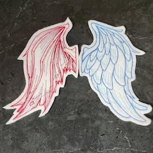 May include: Two embroidered wing patches. One wing is red with detailed stitching, the other is blue with a feathery design. Both are on a white background with a dark grey surface.