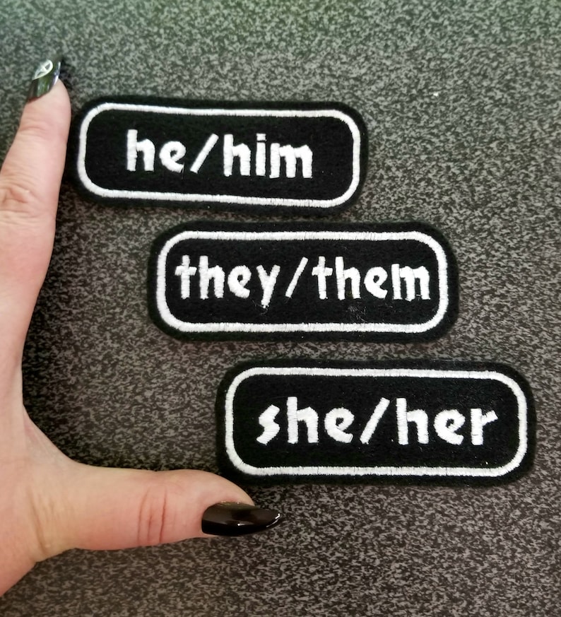 以下が含まれることがあります： 白の刺繍で文字が書かれた黒い布のパッチが3つ。パッチには「he/him」、「they/them」、「she/her」と書いてあります。