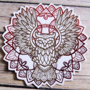 Peut inclure: Patch brodé représentant un hibou brun aux ailes déployées, entouré d'un motif de mandala rouge.