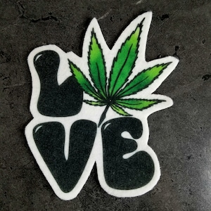 Puede incluir: Un parche de fieltro blanco con la palabra "LOVE" en letras negras delineadas. Una hoja de marihuana verde está detrás de la "O" de la palabra.