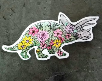 May include: A gray embroidered patch of a triceratops with a floral design. The dinosaur is outlined in black and has pink, yellow, and green flowers.