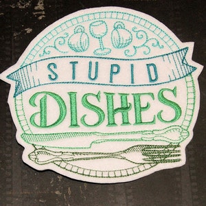 May include: A white embroidered patch with green and blue stitching. The patch is round and features the text "STUPID DISHES" with a fork and knife design.