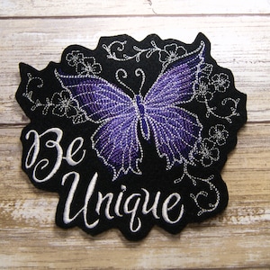 May include: Embroidered iron-on patch with a purple butterfly and white floral designs on a black background. The text "Be Unique" is embroidered in white.