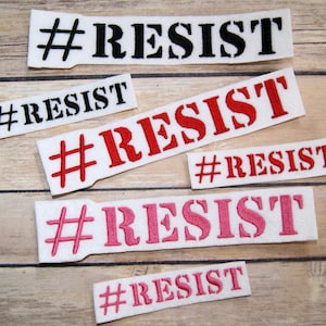 May include: A collection of white fabric patches, each embroidered with the word "RESIST" in black, red, and pink. The patches feature a hashtag symbol and are designed for application to clothing or accessories. Sizes vary.