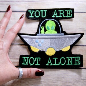 May include: A black felt patch with green embroidered text that reads "You are not alone". A green alien with large eyes is in a gray spaceship with yellow accents.