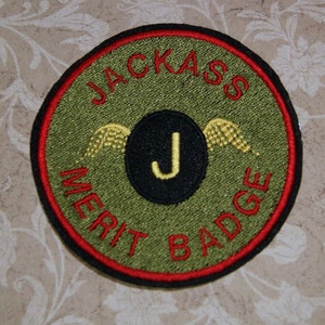 May include: A green and red circular embroidered patch with the text "Jackass Merit Badge" and a black circle with a gold "J" inside. The "J" has wings on either side.