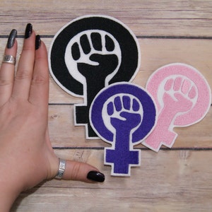 May include: Three iron-on patches in the shape of the female symbol with a raised fist in the centre. The patches are black, purple and pink.