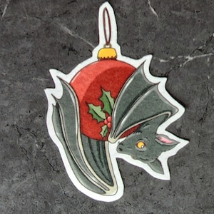 May include: A black bat ornament shaped like a Christmas ornament. The ornament is red with a green holly sprig and red berries. The bat's wings are wrapped around the ornament.