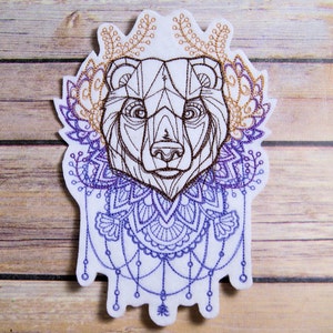 May include: Embroidered patch featuring a geometric bear design with a mandala pattern in shades of purple and gold. The bear's face is outlined in brown thread.