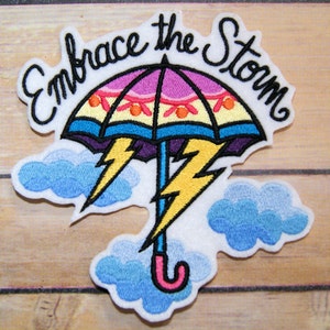 May include: Embroidered iron-on patch with a colorful umbrella, lightning bolts, and clouds. The text "Embrace the Storm" is stitched in a cursive font.