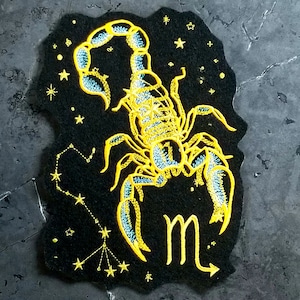 May include: A black felt patch with a yellow embroidered scorpion and the astrological symbol for Scorpio, "m", in yellow. The scorpion is surrounded by small yellow stars.