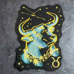 May include: Embroidered iron-on patch featuring a blue and yellow bull with a constellation of stars above it. The bull is wearing a gold chain with stars and the Taurus zodiac symbol is below it.