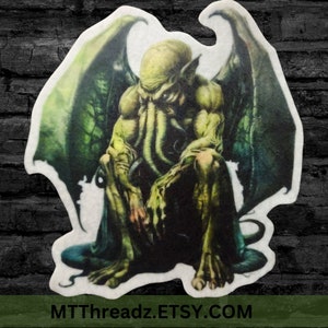 May include: A green, winged, humanoid creature with tentacles sits with its head down. The creature has a large, fleshy head with a pair of large, dark eyes. The creature's wings are large and leathery, and its tentacles are long and thin. The creature is sitting on a white background. MTTThreadz.ETSY.COM is printed at the bottom of the image.