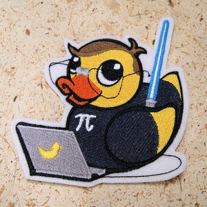 May include: An embroidered iron-on patch featuring a cartoon duck wearing a black shirt with a white 'pi' symbol and holding a blue lightsaber. The duck is wearing glasses and has a laptop in front of it.