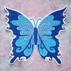 May include: A blue and turquoise embroidered butterfly patch with intricate swirling designs. The patch is outlined in white and has a white backing.
