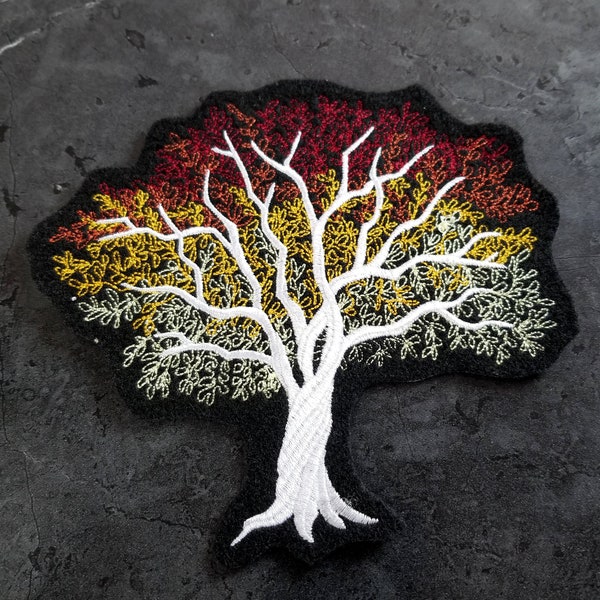 Op de afbeelding: Geborduurde strijkpatch van een witte boom op een zwarte achtergrond. De boom heeft een gradiënt van rode, oranje en gele bladeren.
