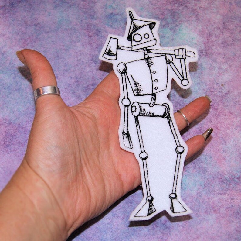 May include: A white felt iron man patch with black embroidery. The iron man is holding an axe and has a hat on his head.