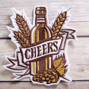 May include: Embroidered iron-on patch with a brown beer bottle, white banner with the word "CHEERS", and gold wheat stalks and hops.