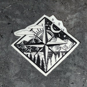 May include: A black and white embroidered patch featuring a compass with a mountain range, sun, moon, and stars. The patch is on a white background.