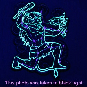 May include: A glow-in-the-dark embroidered patch depicting the constellation Orion. The patch is outlined in blue and features a human figure with a club and a lion's head. The stars are represented by small pink dots.