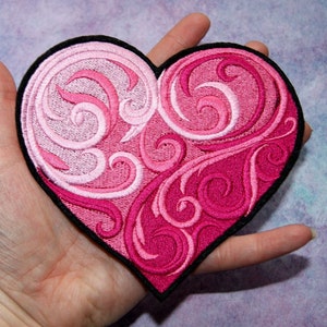 May include: A pink and white heart-shaped embroidered patch with a swirling floral design. The patch has a black border.